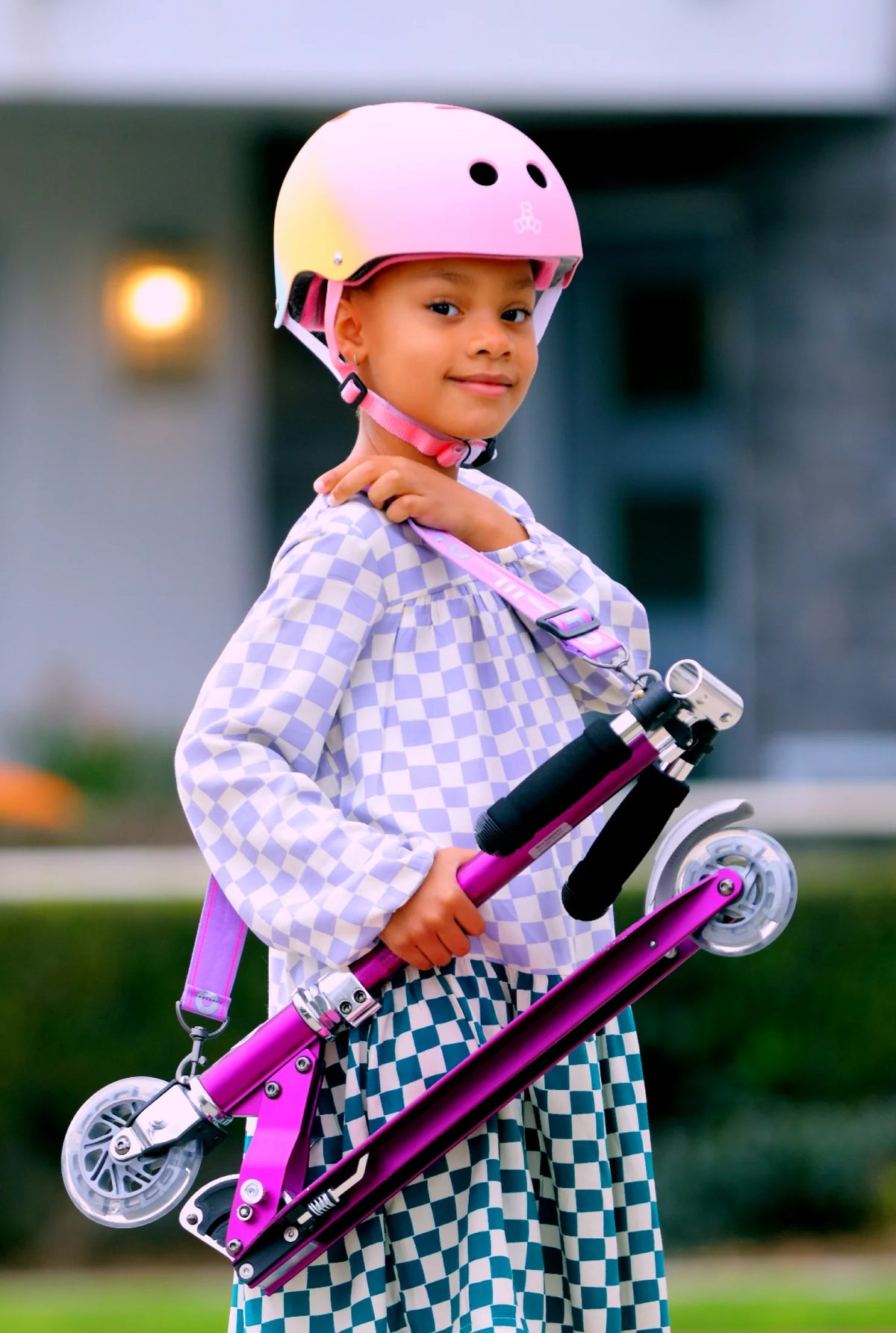 Child wearing a pink helmet and holding a purple scooter outdoors.
