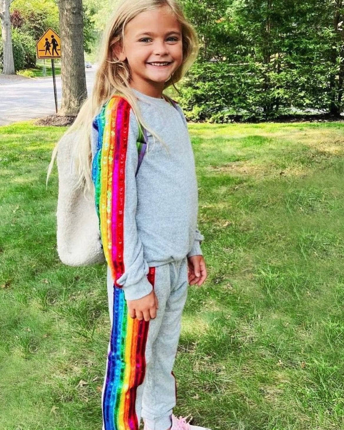 A young girl with blonde hair stands on a grassy lawn wearing a light gray sweatshirt and matching sweatpants, with a large rainbow-striped backpack or shoulder bag. Trees and a road sign are visible in the background.