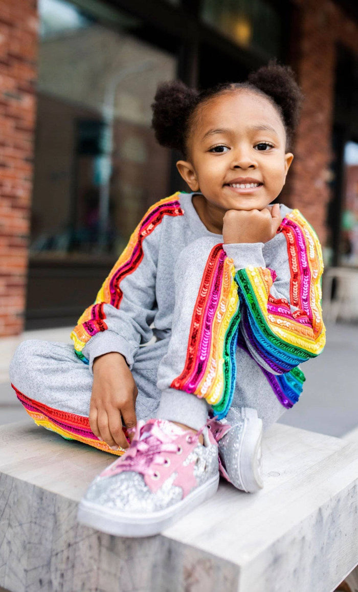A young child sitting on a stone ledge outdoors, wearing a gray tracksuit with bright rainbow sequined stripes and pink glitter sneakers.