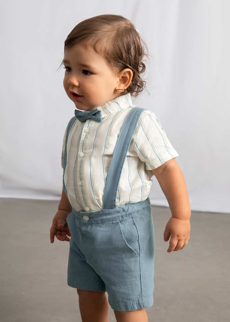 Child wearing a striped shirt, bow tie, and blue suspenders against a plain background