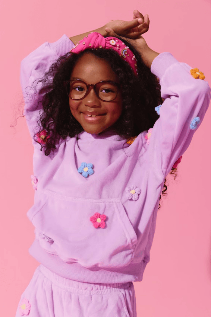 A young girl with curly hair, glasses, and a pink floral headband is wearing a lavender purple tracksuit with colorful 3D flower appliqués, posing against a solid pink background.