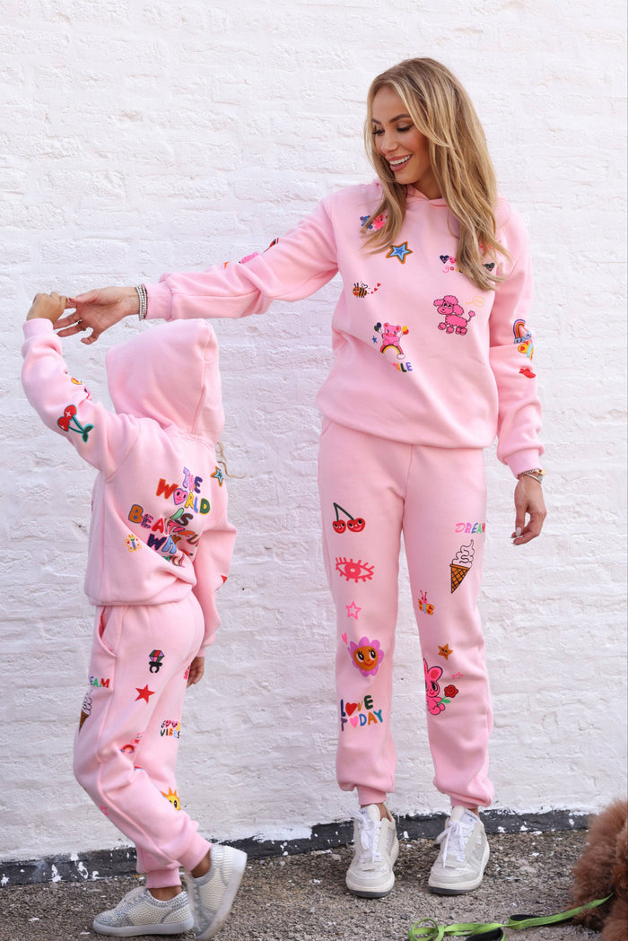 Two people wearing matching pink outfits with colorful designs against a white brick wall.