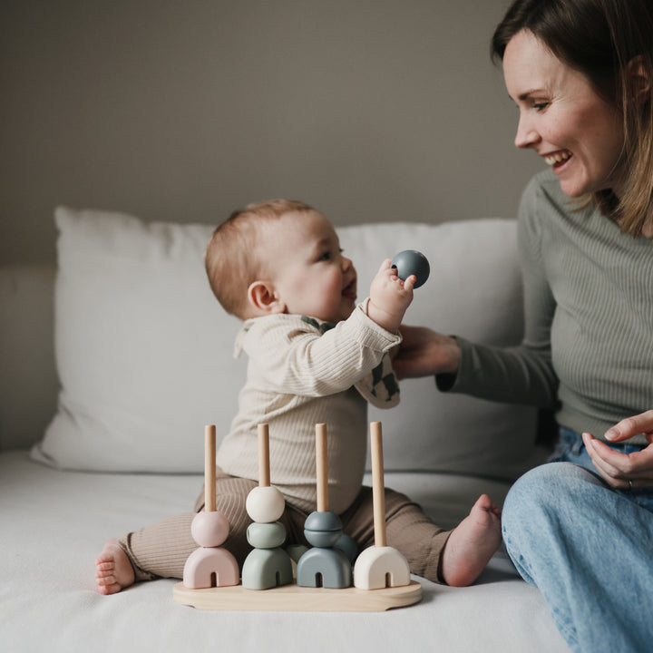 A toddler sitting on a white bed playing with a wooden multi-shape stacker toy while a woman sits beside them smiling.