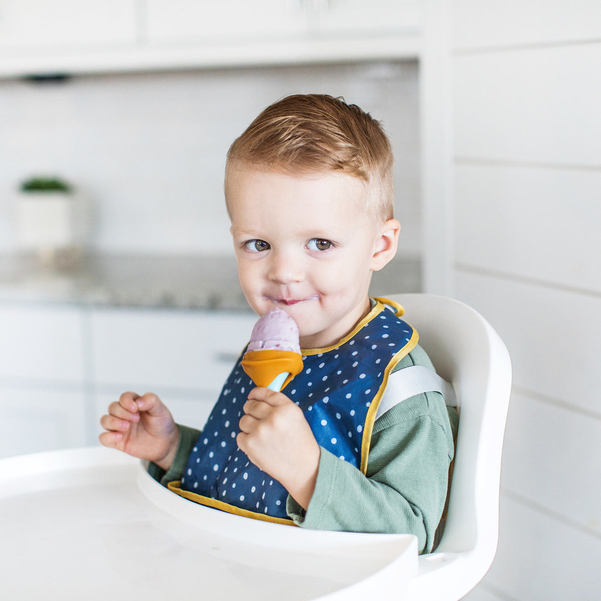Child in a high chair wearing a blue bib, holding an ice cream cone.