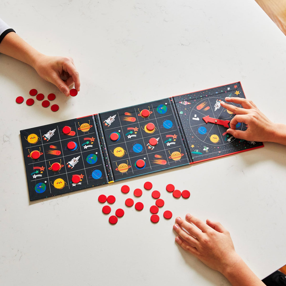 Space Bingo! Magnetic Board Game by Mudpuppy opened layout showing a tri-fold magnetic board with space-themed bingo cards on the left panels and a spinner wheel on the right panel, with someone placing a red magnetic game piece and several other pieces scattered around.