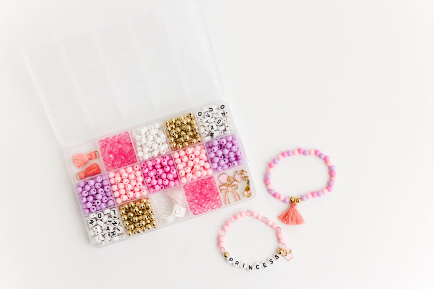 Princess Party DIY Bracelet Kit featuring a compartment storage box filled with colorful beads, alphabet letter beads, and charms next to two completed pink and gold beaded bracelets, including one with a tassel charm and one spelling "PRINCESS".