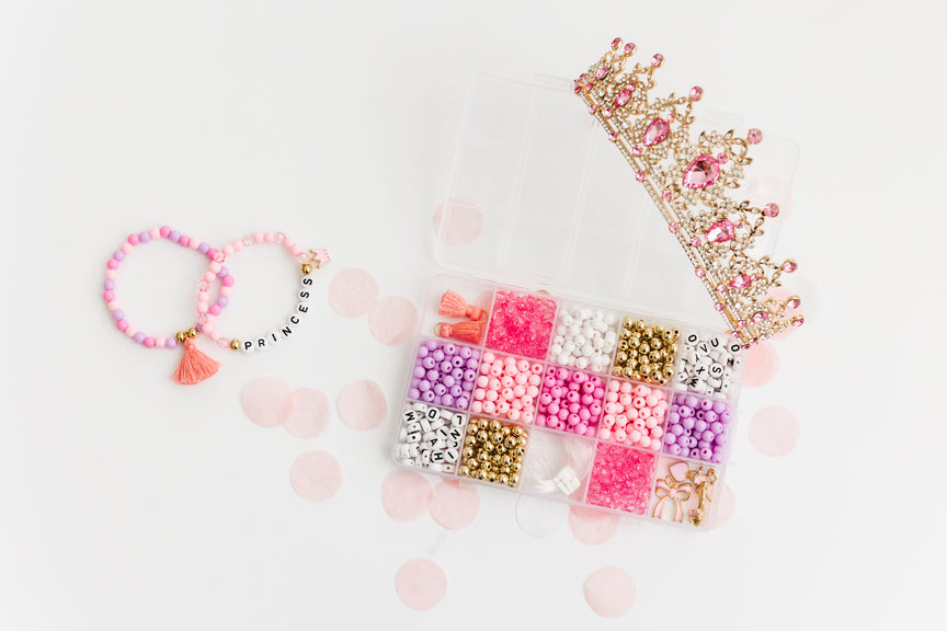 Princess Party DIY Bracelet Kit featuring an open compartment storage box filled with colorful beads, alphabet letter beads, and charms, accompanied by two completed beaded bracelets (one with a tassel) and a decorative gold tiara crown on a light background.