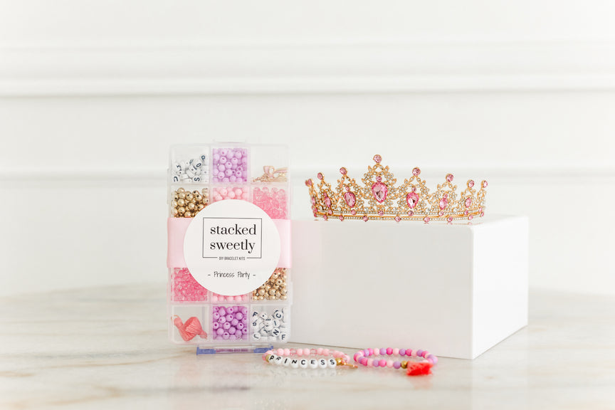 Princess Party DIY Bracelet Kit featuring a compartment storage box filled with colorful beads, alphabet letter beads, and charms next to two completed pink and gold beaded bracelets, accompanied by a decorative pink-jeweled gold tiara crown.