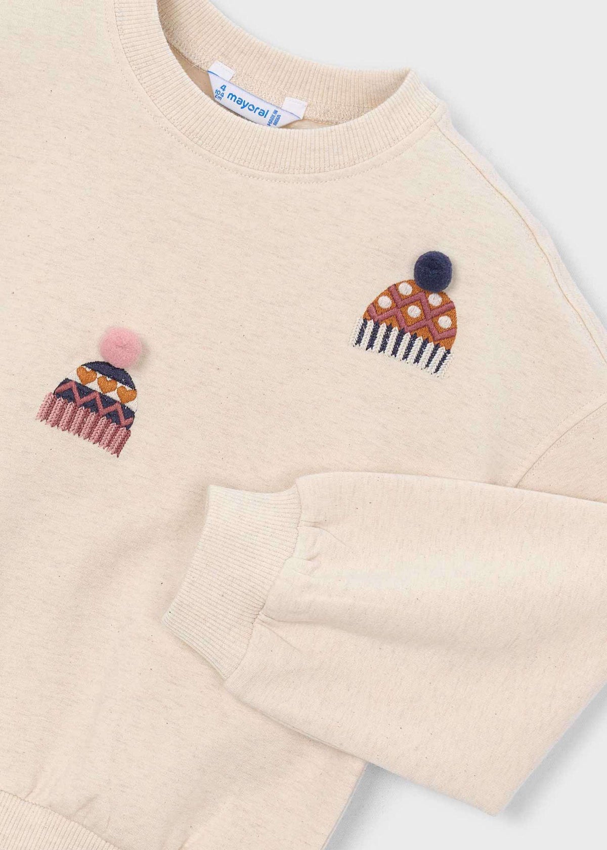 A close-up of a cream-colored crewneck sweatshirt featuring two embroidered knit hat motifs on the chest. The left chest has a pink and orange patterned hat with a pink pom-pom, and the right chest has a brown and orange polka-dot hat with a dark blue pom-pom.