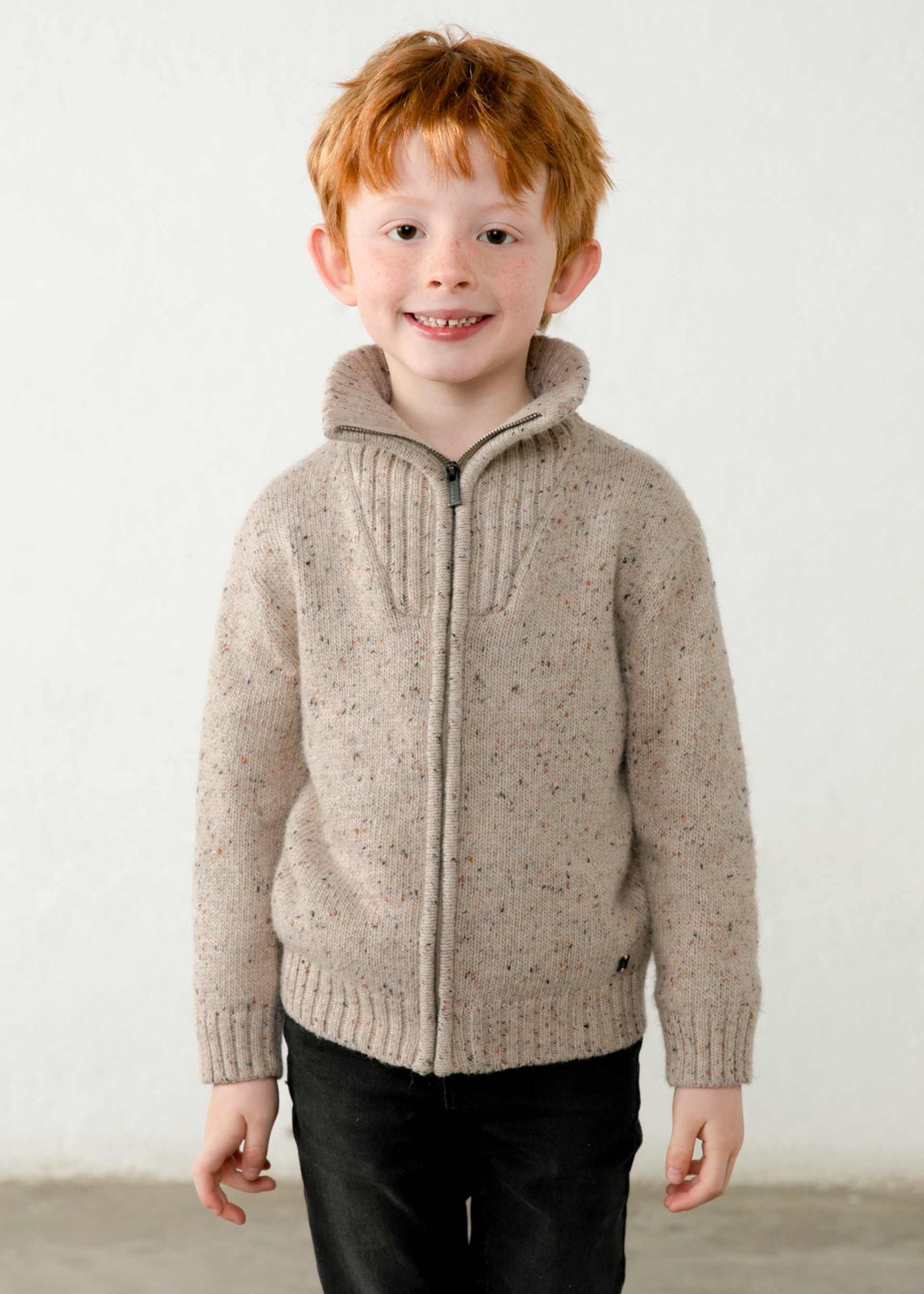 A young boy with red hair wearing a beige speckled knit zip-up sweater with a mock neck collar and black pants, standing against a plain off-white wall.