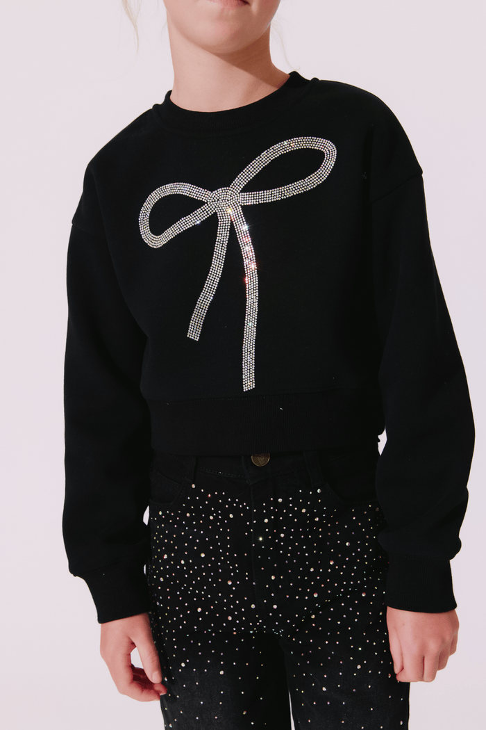 A person wearing a black cropped sweatshirt with a large sparkling silver bow graphic on the front, paired with black pants covered in small silver rhinestones, against a pale pink background.