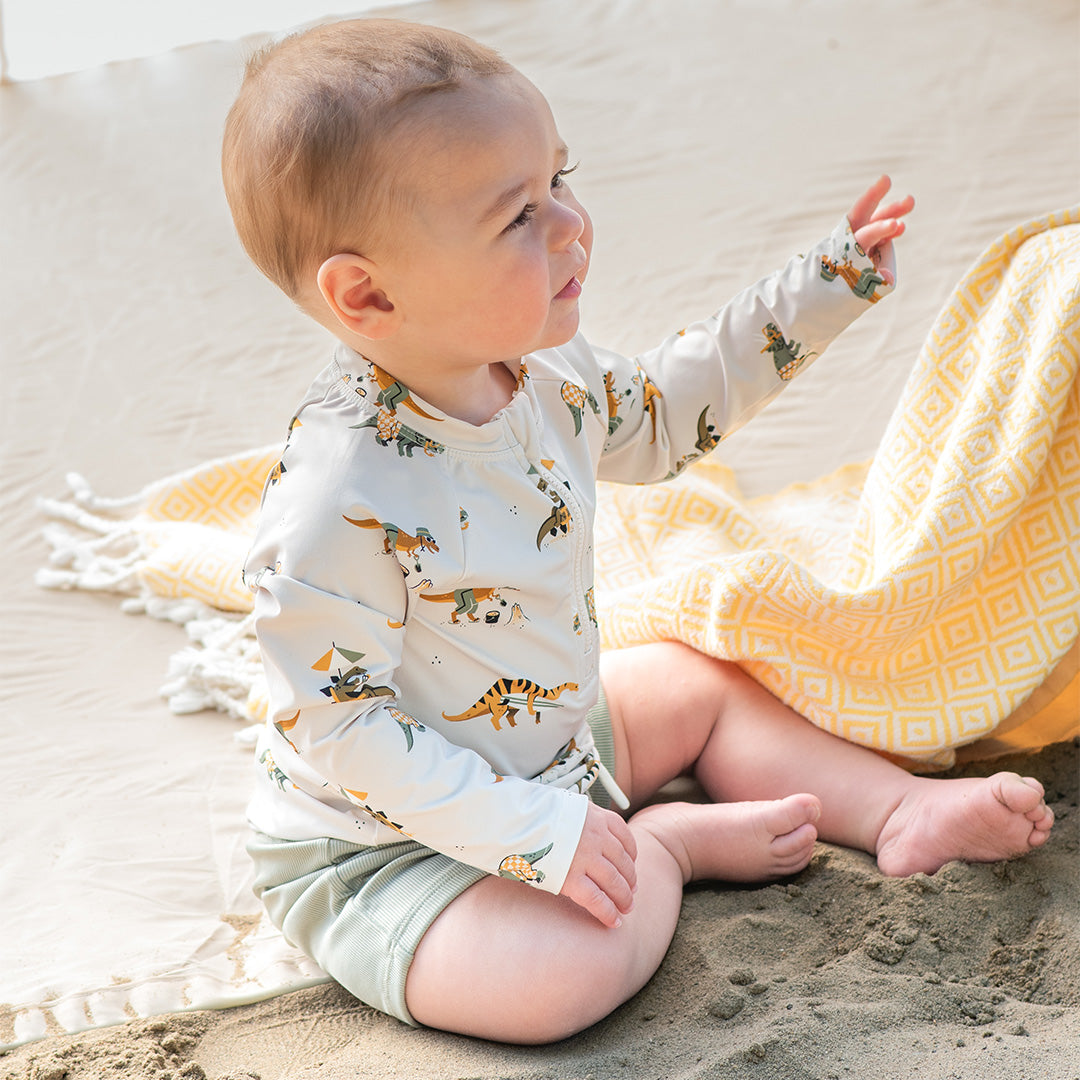 Baby sitting on sand wearing a dinosaur-patterned shirt and shorts