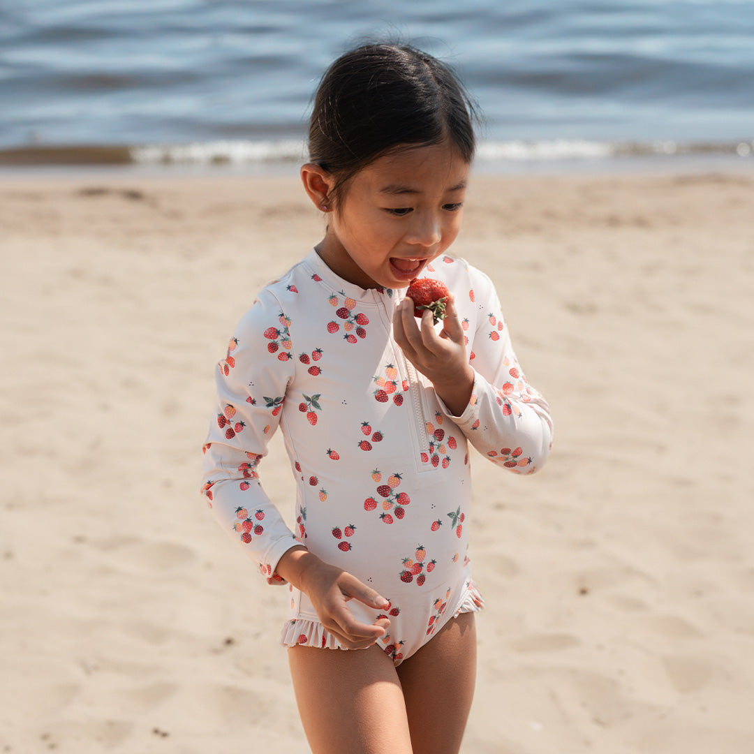 Child in a floral swimsuit eating a strawberry on a beach