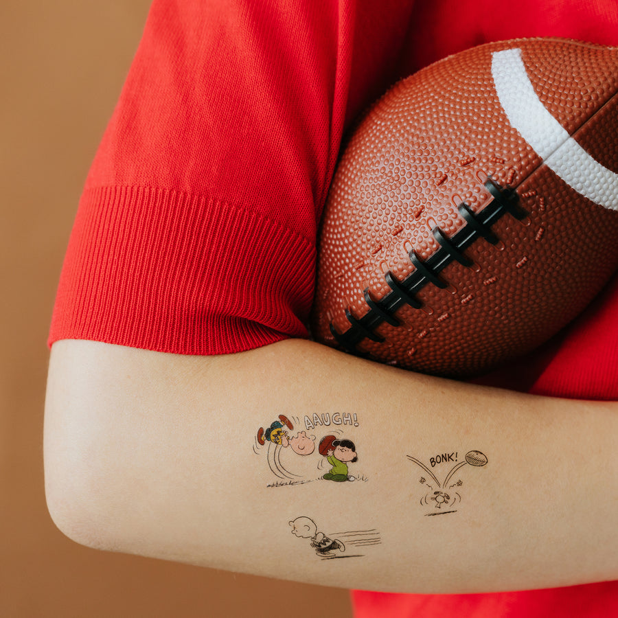 Person holding a football with tattoos of cartoon characters on their arm against a brown background