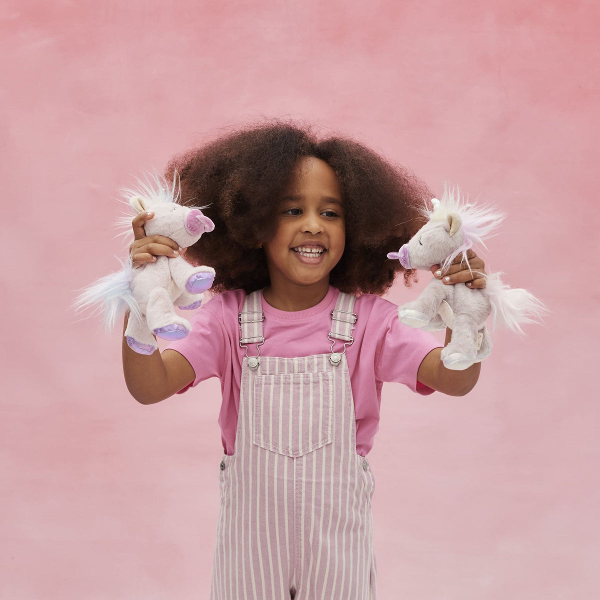 Child holding two stuffed unicorns against a pink background