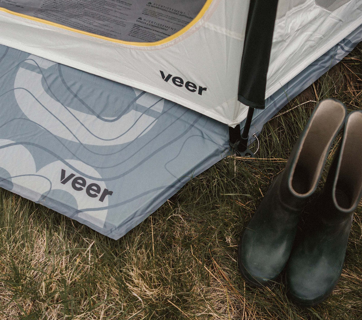 Camping setup with 'veer' branded tent and sleeping pad on grass, accompanied by green rubber boots.