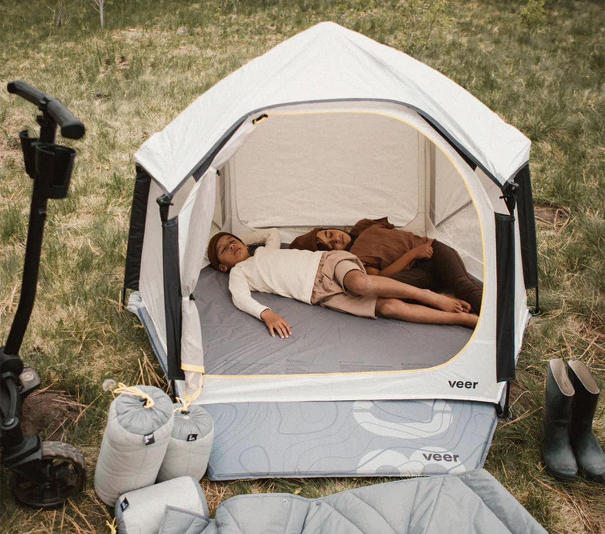 Two people lying inside a portable camping pod with 'veer' branding on a grassy outdoor setting.