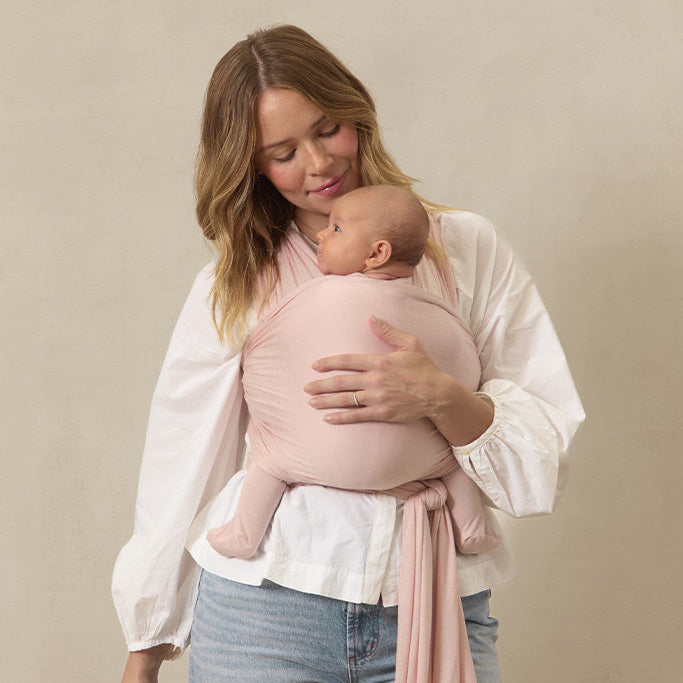 Woman holding a baby wrapped in a pink sling against a beige background
