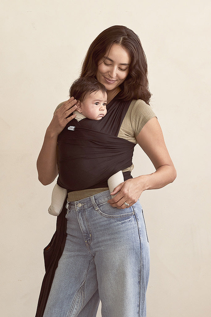 Woman holding a baby in a black baby carrier against a beige background