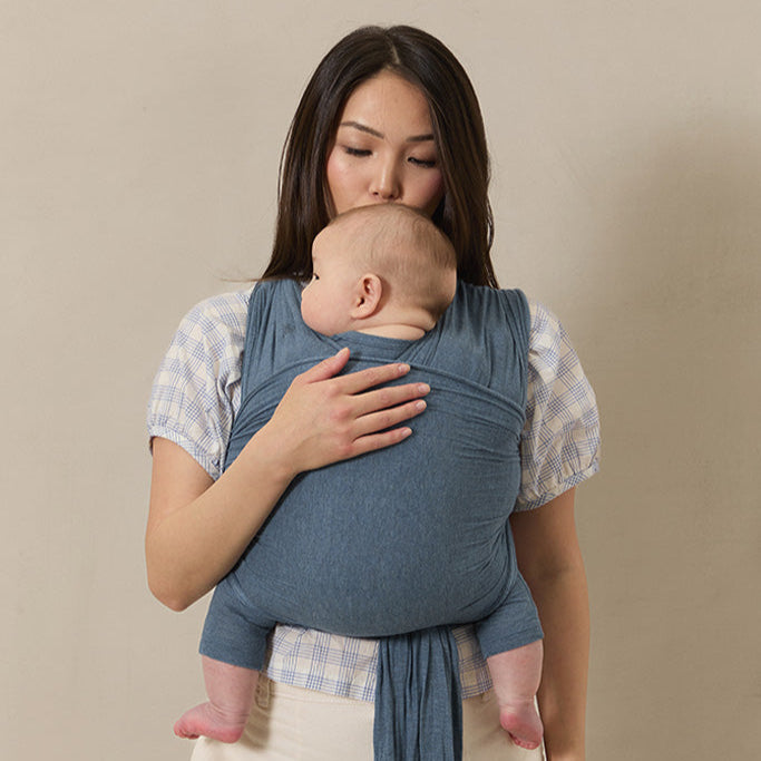 Woman holding a baby in a blue sling against a beige background