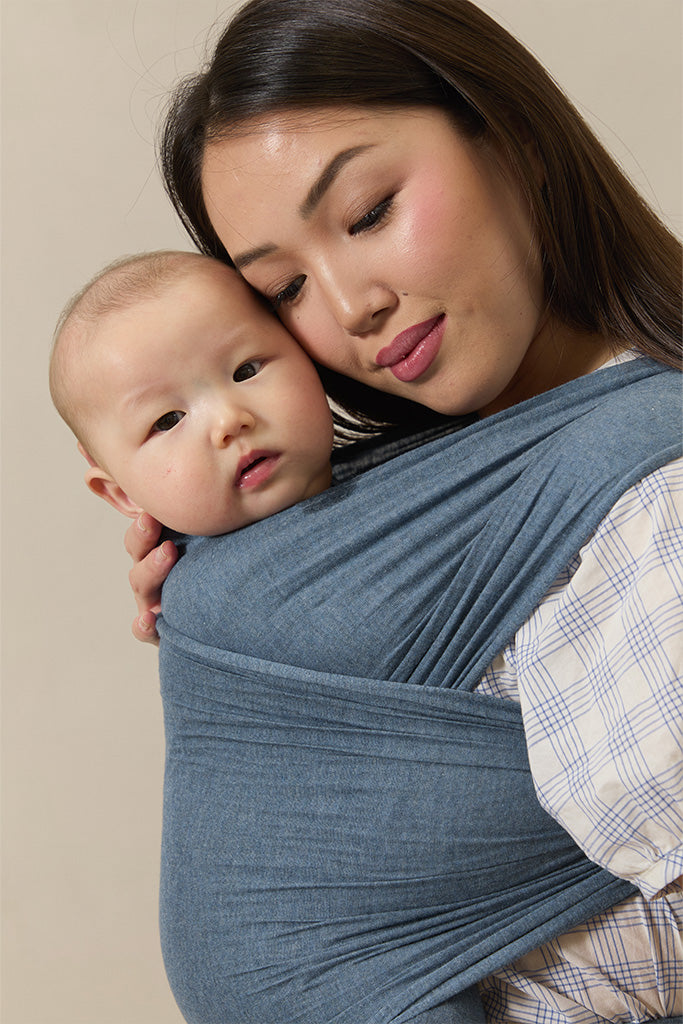 Woman holding a baby wrapped in a blue sling against a beige background