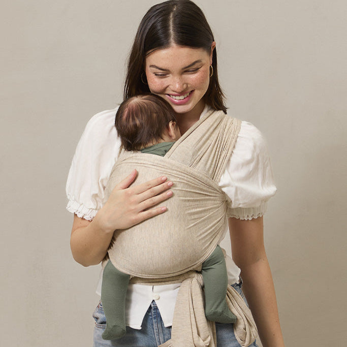 Woman holding a baby in a beige baby carrier against a plain background