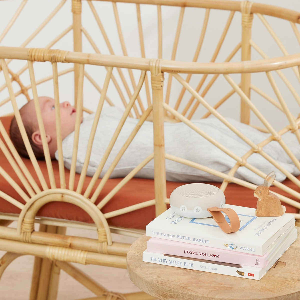 Baby lying in a rattan crib with a small table next to it featuring books and a toy.