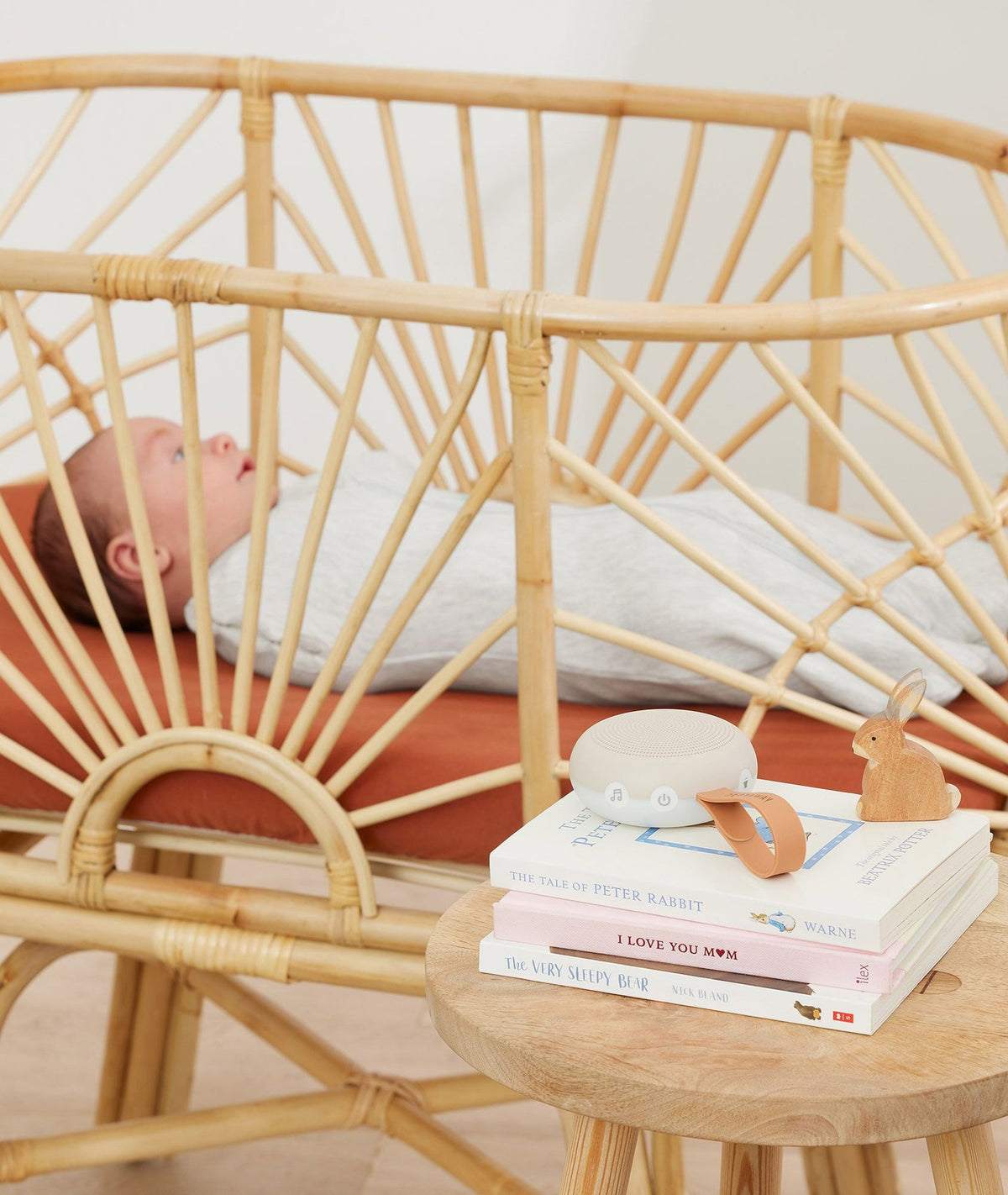 Baby lying in a rattan crib with a small table next to it featuring books and a toy.