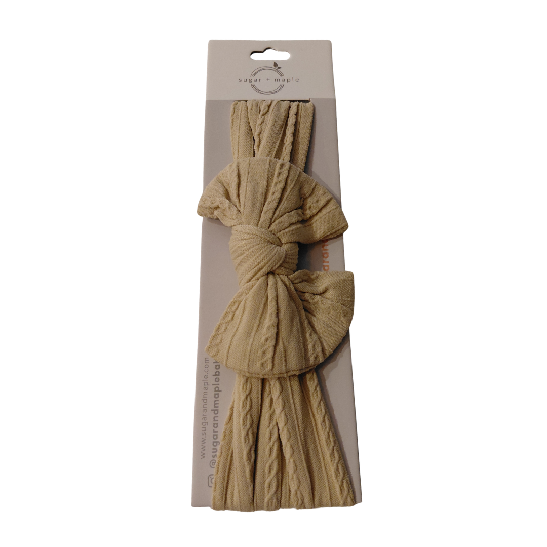 A beige cable-knit hair bow accessory in its original retail packaging, featuring a knotted bow with matching knitted tails on a cardboard backing.