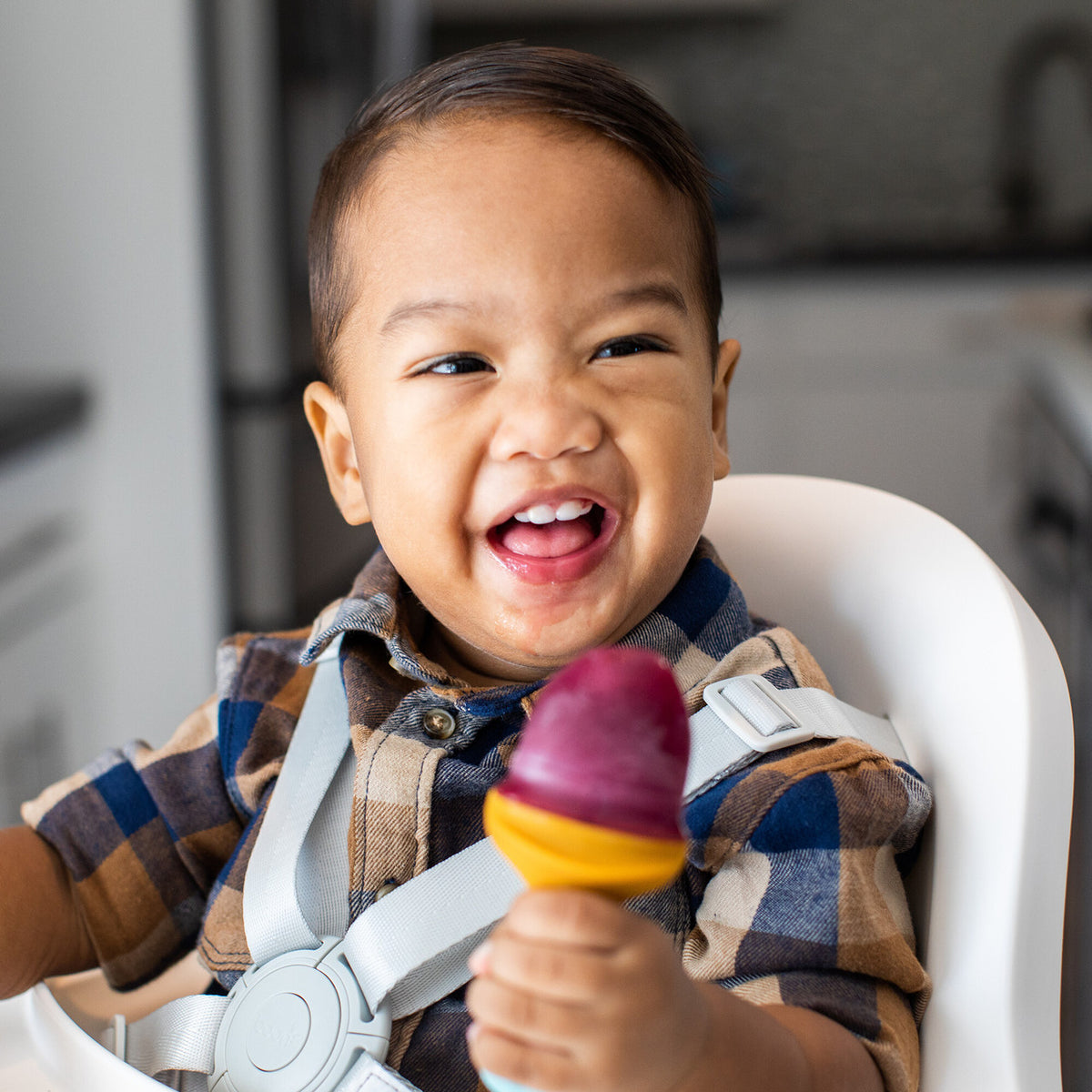 A smiling young child wearing a brown and blue plaid shirt sits in a white high chair, holding a colorful popsicle with a purple top and yellow cone.