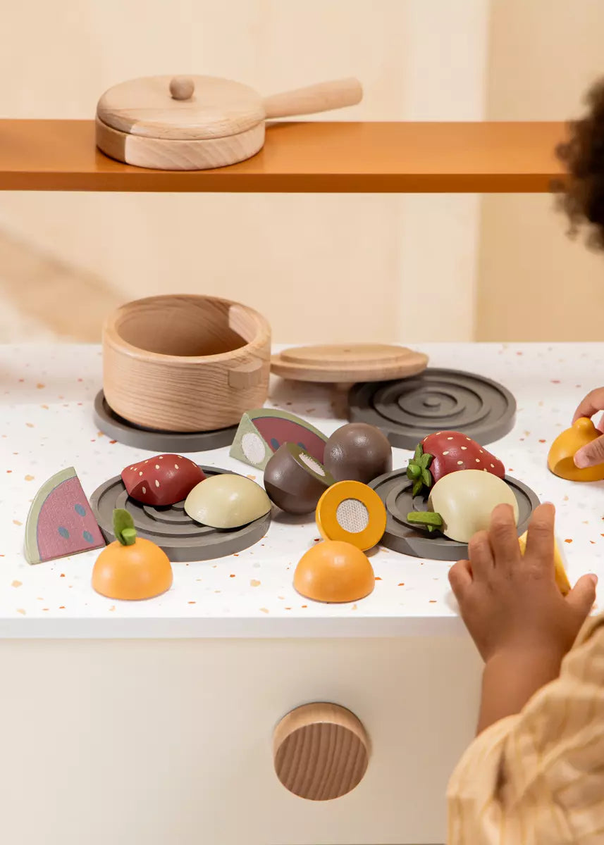 Children's play kitchen set with wooden utensils and toy food on a white surface.