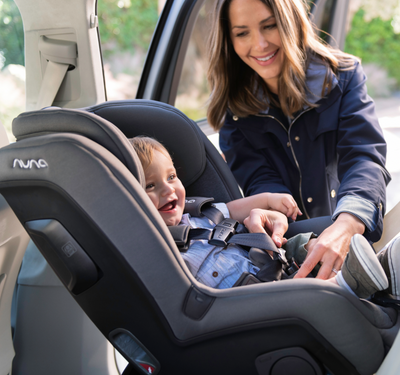 Photo of a mom buckling her smiling child into a black Nuna carseat.