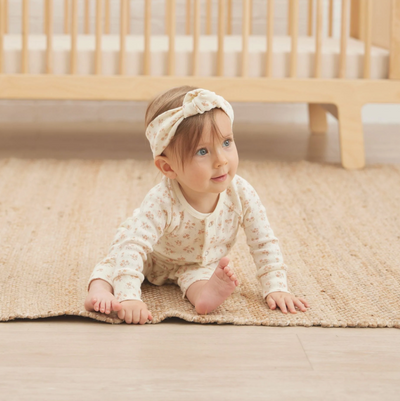 Baby sitting on a rug with a crib in the background while wearing a matching headband and long sleeve patterned romper.