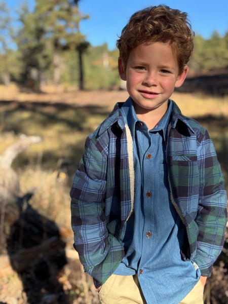 Young boy wearing a blue plaid shirt and jacket outdoors with trees in the background