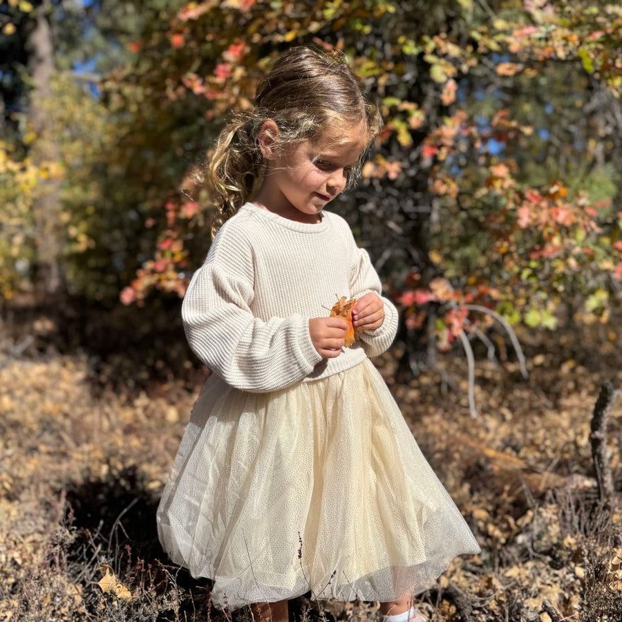 A young girl stands outdoors in a wooded area wearing a cream ribbed long-sleeve top and a champagne tulle skirt, holding a small orange leaf.