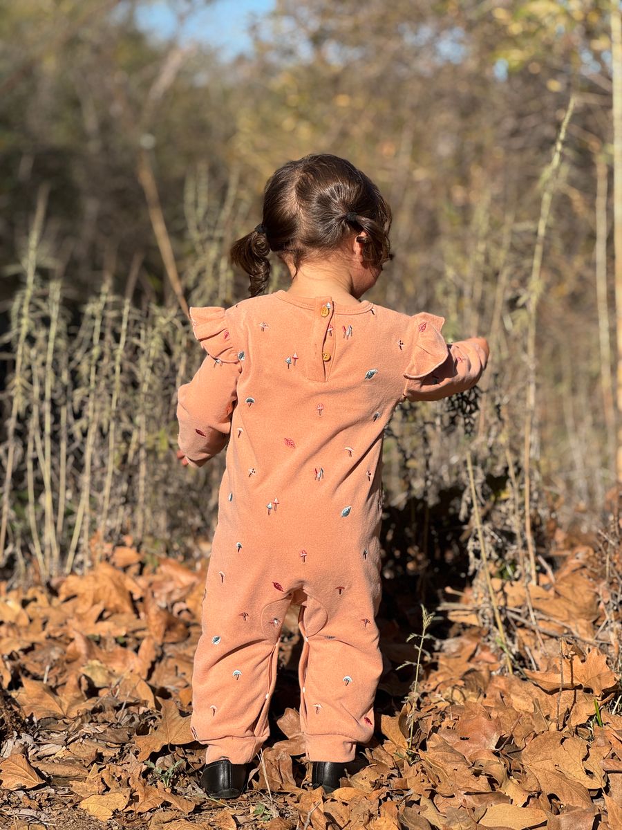 A toddler viewed from behind wearing a peach-colored ruffle-shoulder jumpsuit with small colorful embroidered details, standing outdoors on a bed of dry autumn leaves.