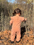 A toddler viewed from behind wearing a peach-colored ruffle-shoulder jumpsuit with small colorful embroidered details, standing outdoors on a bed of dry autumn leaves.