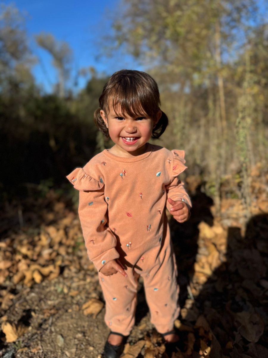 A smiling toddler standing outdoors in a wooded area, wearing a peach-colored long-sleeve romper with small scattered animal prints.