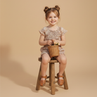Young girl sitting on a wooden stool holding a wicker basket with flowers against a beige background