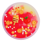 A round, translucent pink and yellow gel toy filled with colorful decorative elements, including honeycomb shapes, white dots, a small red apple with a green leaf, and a striped honey pot decoration, shown on a plain white background.
