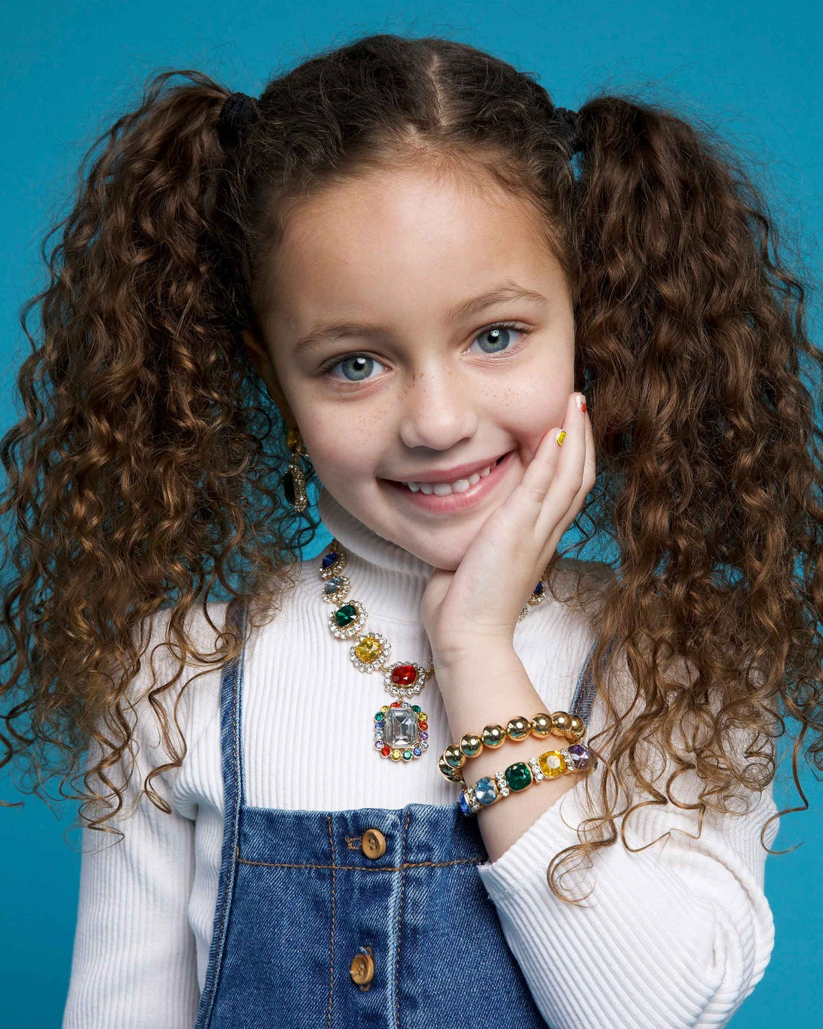 Young girl with curly hair in pigtails wearing colorful jewelry against a blue background