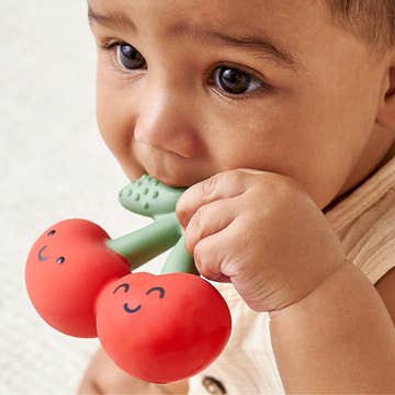 Baby holding a red and green teething toy with a smiley face on a light background