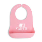 A pink silicone baby bib with a wide chin guard and front pocket, featuring the text 'MINI SWIFTIE' and 'BELLA TURNUP' printed in white. The bib is shown on a clean white background with a soft drop shadow.