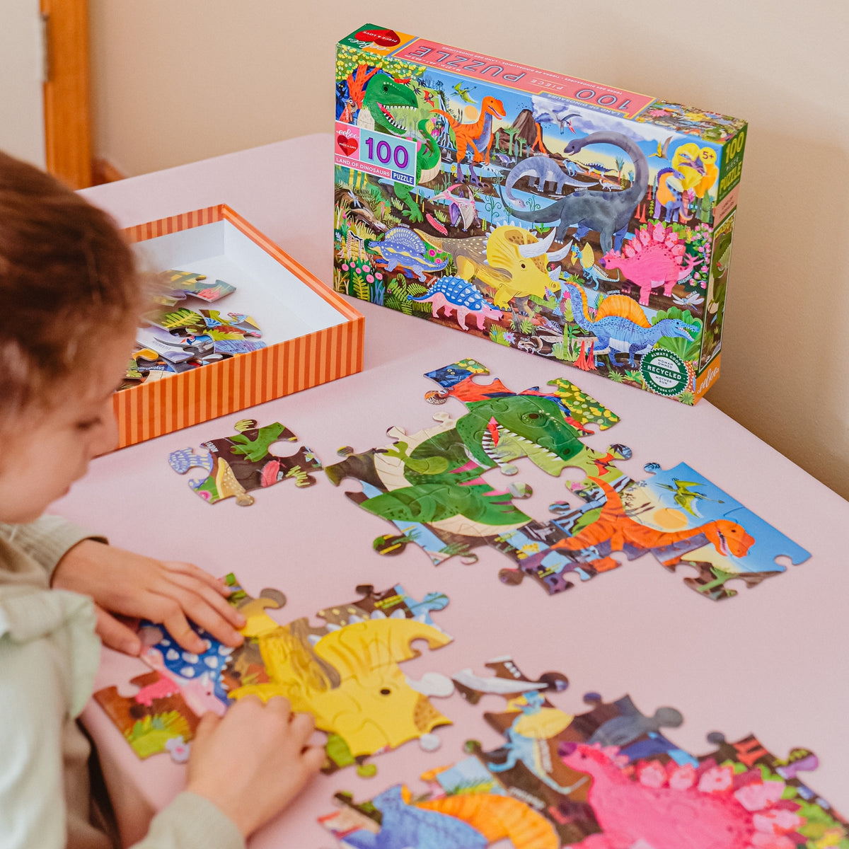 Child playing with a dinosaur-themed puzzle on a table next to the puzzle box.