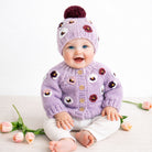 Baby wearing a purple floral cardigan and matching hat with a pom-pom, sitting on a white surface with flowers.
