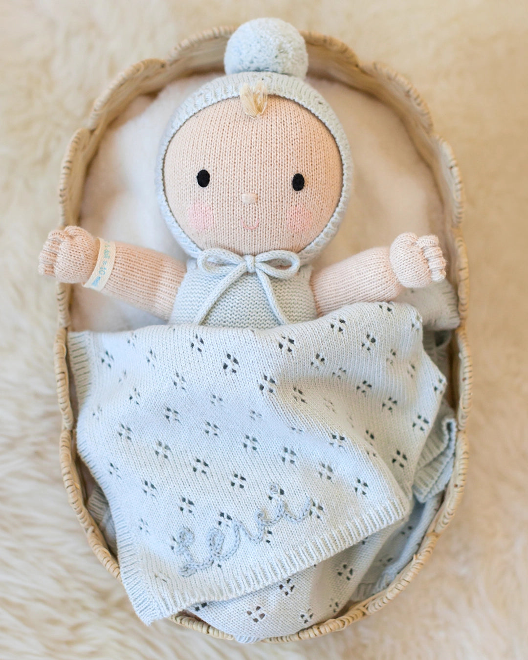 A handmade knitted baby doll with a pale peach face and hands, wearing a light blue bonnet and bodysuit, nestled inside a woven straw basket with a matching knitted blanket that says 'Lola'.