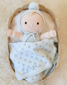 A handmade knitted baby doll with a pale peach face and hands, wearing a light blue bonnet and bodysuit, nestled inside a woven straw basket with a matching knitted blanket that says 'Lola'.