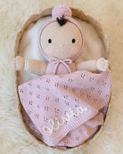 A handmade knitted doll with a pink bonnet and matching blanket sits inside a woven basket on a soft cream-colored rug. The blanket is embroidered with the name 'Olivia' in white script.
