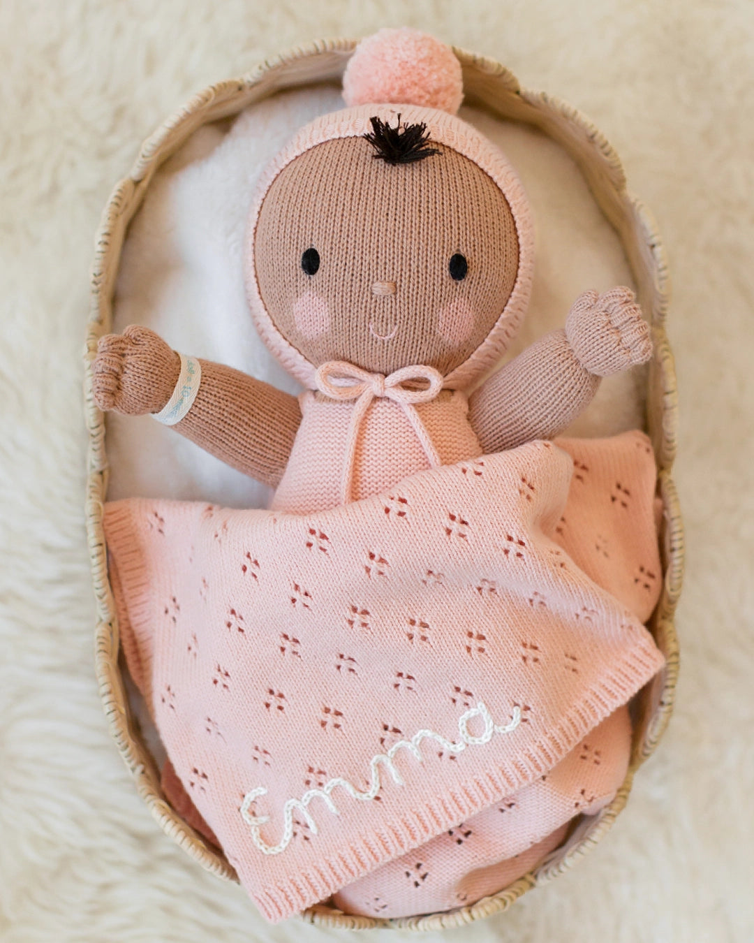 A handmade knit baby doll wrapped in a pink blanket with the name 'Emma' embroidered in white, resting inside a woven basket on a soft cream background.