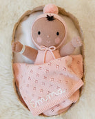 A handmade knit baby doll wrapped in a pink blanket with the name 'Emma' embroidered in white, resting inside a woven basket on a soft cream background.